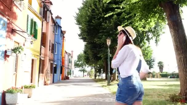 Venetië, Burano, Italië - 7 juli 2018: jonge vrouw, meisje in zonnebril, hoed, broek, praten over de telefoon, glimlachend. Burano eiland met multicolor huizen, in de buurt van Venetië. — Stockvideo