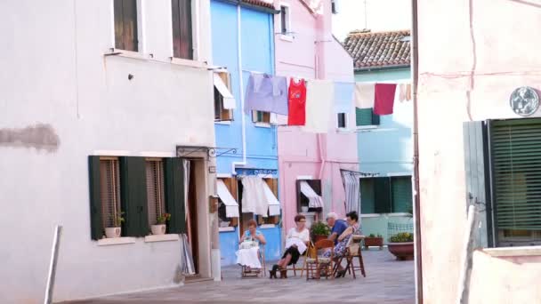 Venedik, Burano, İtalya - 7 Temmuz 2018: atmosferik Caddesi'nde Burano adası. Yaşlı insanlar sokakta oturup, nakış, sohbet. evleri arasında üst çamaşırları kurutulur. — Stok video