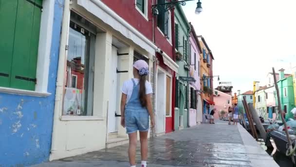 VENICE, BURANO, ITÁLIA - JULHO 7, 2018: menina bonita em macacões jeans azuis, shorts e boné, caminha pelas ruas, ao longo do pequeno canal de Burano. verão dia quente . — Vídeo de Stock