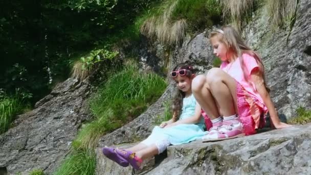 Zwei hübsche Mädchen, Kinder, sitzen auf einem Felsvorsprung, ein wunderschöner orangefarbener Bergschmetterling wirbelt und fliegt in Sandalen über ein Kinderbein. Schmetterling sitzt auf dem Fuß. Heißer Sommertag — Stockvideo