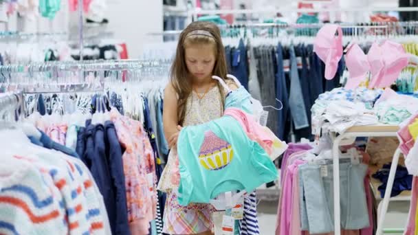 Compras tediosas nas lojas. criança cansada, triste, menina, com um monte de coisas em suas mãos, esperando os pais na loja . — Vídeo de Stock