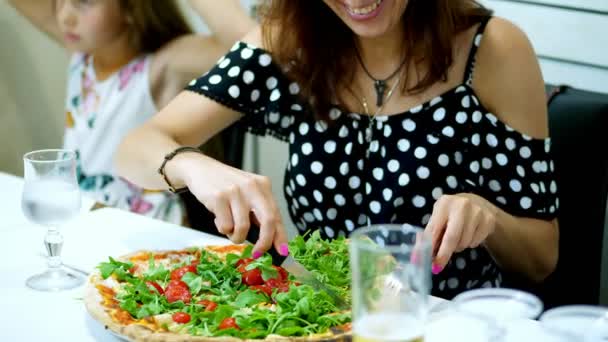 In a pizza, a woman cuts a large, juicy, hot pizza with greens, arugula and cherry tomatoes. pizza to pieces with a knife and fork. The woman is pretty smiling. — Stock Video