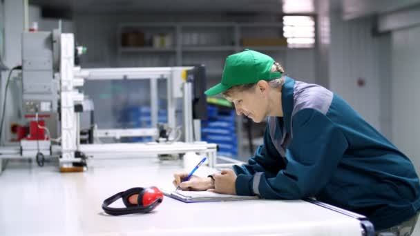 Ältere Frau, Angestellte, Arbeiterin in einem Unternehmen, Fabrik, füllt ein Dienstleistungstagebuch, Aufzeichnungsbuch, arbeitender industrieller Hintergrund. — Stockvideo