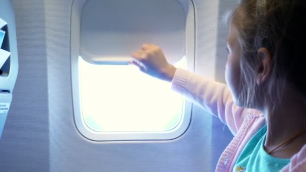 Close-up. jongen meisje werpt de patrijspoort gordijn in de cabine van de vliegtuigen, vanaf daar schijnt een helder licht. meisje op zoek door vliegtuigen raam hemel en wolken en landschap hieronder bekijken — Stockvideo