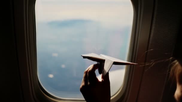 Gros plan. Silhouette d'une main d'enfant avec un petit avion en papier sur le fond de la fenêtre de l'avion. Enfant assis près de la fenêtre de l'avion et jouant avec un petit avion en papier. pendant le vol en avion — Video