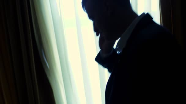 Za šera, ve tmě, naproti oknu. pohledný muž, podnikatel v bílou košili a modré sako drží smartphone, mluvit na mobilním telefonu, doma, ve své ložnici — Stock video