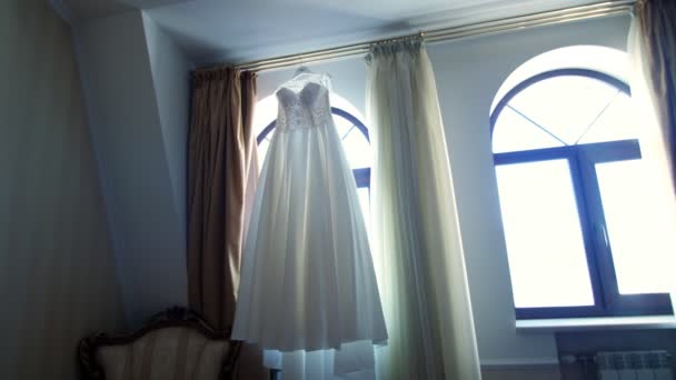 Около окна, в комнате, белое свадебное платье висит на оконных карнизах — стоковое видео