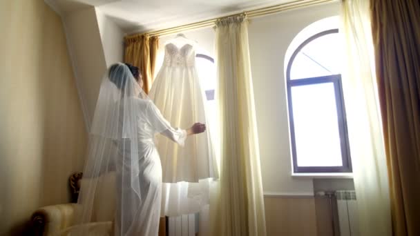 附近的窗口, 在房间里, 白色的婚纱挂在窗口屋檐上。新娘, 一个穿着白色长袍的漂亮女孩, 一件浴衣, 正在考虑她的婚纱, 为婚礼做准备. — 图库视频影像