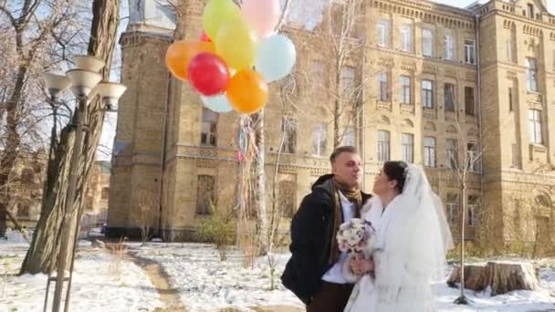 Winterhochzeit, glückliches, lachendes Brautpaar mit bunten bunten Luftballons. sonniger, verschneiter Tag, die antike Architektur der Stadt. — Stockvideo