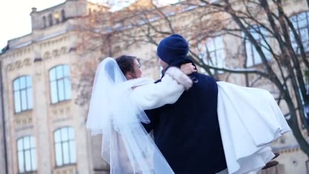 Winterhochzeit. Brautpaar in Brautkleidern. Der Bräutigam hält die Braut im Arm und dreht sich. Sie sind glücklich und lächeln sich an. Hintergrund der antiken Architektur, schneebedeckter Park, — Stockvideo