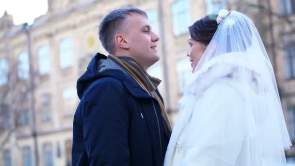 Vinter bröllop. nygift par i bröllopsklänningar. brudgummen kysser försiktigt bruden på pannan, kram. de är glada, ler mot varandra. bakgrund av antik arkitektur, snötäckta park, — Stockvideo