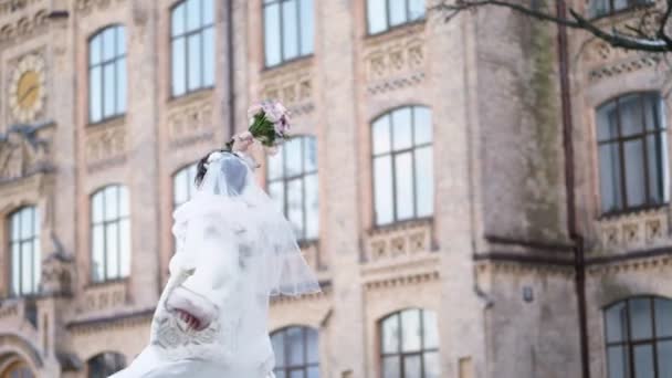 Retrato de uma bela noiva, em um vestido branco, véu e casaco de pele. noiva está girando, e sorrindo feliz, segurando um buquê de casamento. contra o pano de fundo da arquitetura antiga, casamento de inverno . — Vídeo de Stock