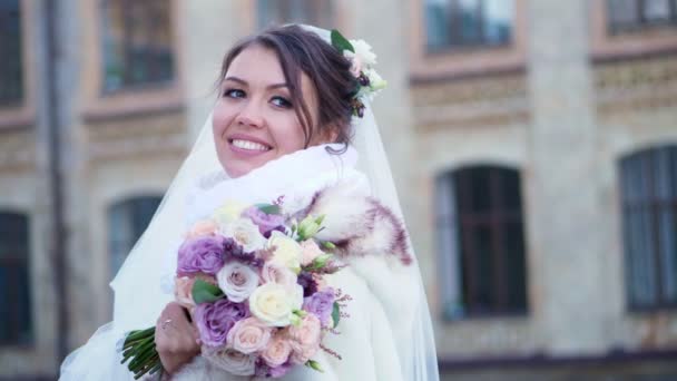 Retrato de uma bela noiva, em um vestido branco, véu e casaco de pele. noiva está sorrindo feliz, segurando um buquê de casamento. contra o pano de fundo da arquitetura antiga, casamento de inverno . — Vídeo de Stock