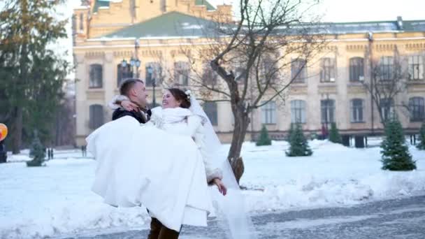 冬の結婚式します。ウェディング ドレスで新婚カップル。新郎は彼の腕の回転の中で花嫁を保持します。彼らは満足して、お互いに笑みを浮かべています。古代建築、雪に覆われた公園の背景, — ストック動画