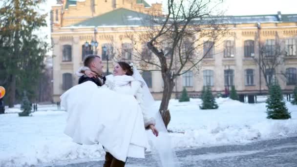 Zimowy ślub. Newlywed para w suknie ślubne. Pan młody posiada Panna Młoda w jego ramionach, spinning. są szczęśliwi, uśmiechając się do siebie. tle architektury starożytnej, pokrytych śniegiem park, — Wideo stockowe