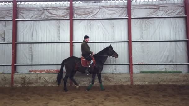 En el hangar especial, un joven discapacitado aprende a montar un caballo negro, pura sangre, hipoterapia. El hombre tiene un miembro artificial en lugar de su pierna derecha. concepto de rehabilitación de discapacitados con — Vídeo de stock