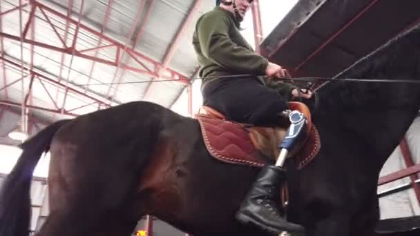 在特殊的机库里, 一个年轻的残疾人学会了骑一匹黑色的纯种马, 进行嬉皮士治疗。人有假肢而不是右腿。残疾人康复的概念 — 图库视频影像
