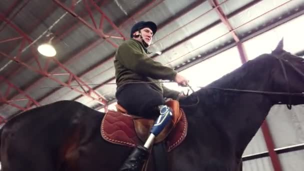 In speciale hangar leert een jonge gehandicapte man paardrijden zwart, volbloed, hippotherapie. man heeft een kunstmatige ledematen in plaats van zijn rechterbeen. concept van de revalidatie van mensen met een handicap met — Stockvideo