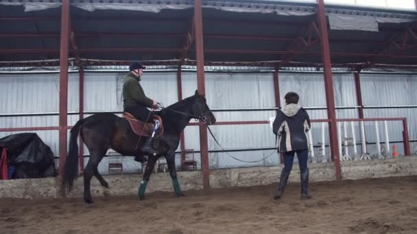 In apposito hangar, un giovane disabile impara a cavalcare un cavallo con insegnante di stretta supervisione, ippoterapia. l'uomo ha un arto artificiale invece della gamba destra. riabilitazione di disabili con animali . — Video Stock