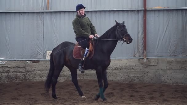 En el hangar especial, un joven discapacitado aprende a montar un caballo negro, pura sangre, hipoterapia. El hombre tiene un miembro artificial en lugar de su pierna derecha. concepto de rehabilitación de discapacitados con — Vídeo de stock