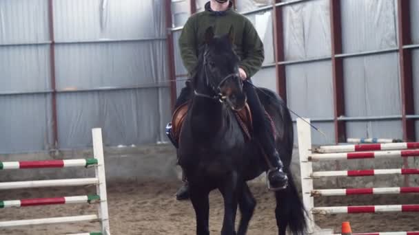 En el hangar especial, un joven discapacitado aprende a montar un caballo negro, pura sangre, hipoterapia. El hombre tiene un miembro artificial en lugar de su pierna derecha. concepto de rehabilitación de discapacitados con — Vídeos de Stock