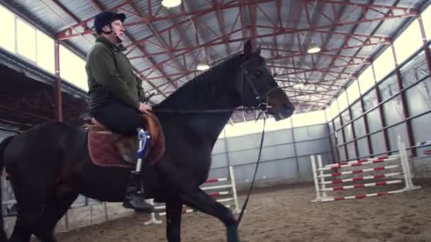 In un hangar speciale, un giovane disabile impara a cavalcare un cavallo nero, purosangue, ippoterapia. l'uomo ha un arto artificiale invece della gamba destra. concetto di riabilitazione dei disabili con — Video Stock