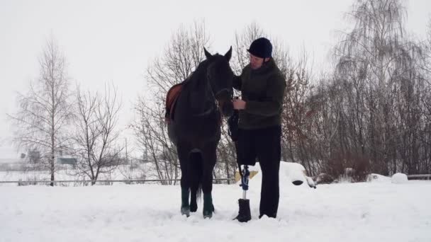 Zimní, zakázaný člověk stojí u Černého koně na zasněženou plání. muž tahy tlamy koně. člověk má protézu místo pravou nohu. on se učí jezdit na koni, hipoterapie. rehabilitace — Stock video