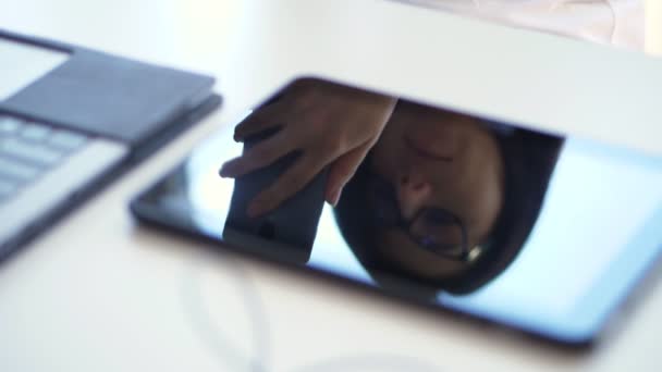 In Großaufnahme, auf einem weißen Desktop, neben einem Laptop, spiegelt sich im Tablet-Bildschirm das Gesicht einer Geschäftsfrau mit Brille. Sie schaut etwas in einem Mobiltelefon. Telefonieren auf dem Handy. — Stockvideo
