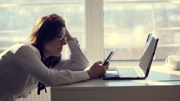 Μια νεαρή γυναίκα υπάλληλος γραφείο εργαζόμενος μεταβεί κοινωνικά δίκτυα σε ένα κινητό τηλέφωνο, κατά τη διάρκεια της ημέρας εργασίας, σε ένα γραφείο. ένα σύντομο διάλειμμα, ανάπαυση στο έργο. πιθανή έννοια αδίστακτοι και τεμπέλης υπαλλήλου ή — Αρχείο Βίντεο