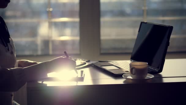 Σκοτεινή σιλουέτα του επιχειρηματίας, closeup των χεριών. γράφει κάτι σε δισκίο, επόμενο είναι ένα φορητό υπολογιστή στο τραπέζι. Παράθυρο θολή φόντο, μια ακτίνα φωτός αντανακλάται στην οθόνη του tablet — Αρχείο Βίντεο