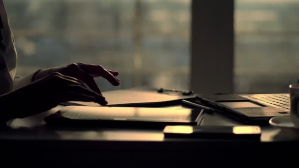 Closeup, dark silhouette of female hands. she is typing something in tablet, next is a cup, mobile, laptop on table. Blurred window background , a ray of light is reflected in the tablet screen — Stock Video