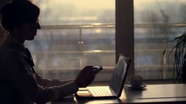 Genç odaklı iş kadını gözlük, karanlık siluet bir şey onun mobil olarak yazarak, sonraki tablo üstünde laptop. gün batımında, ışınları içinde bir büyük ofis pencere arka planı