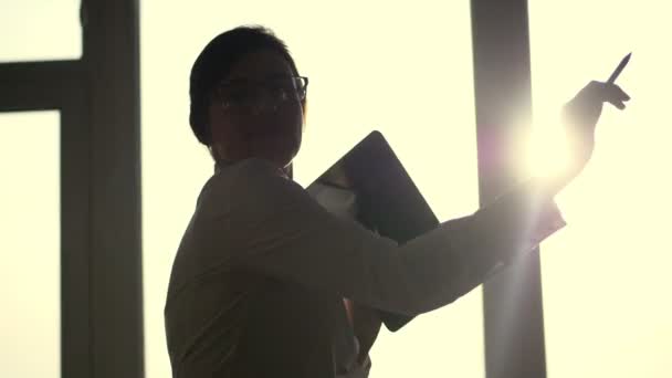 Temná silueta podnikatelka, nosí brýle, tance s tabletem v ruce, na pozadí velké kanceláře okna, při západu slunce, v paprscích světla. obchodní žena slaví — Stock video
