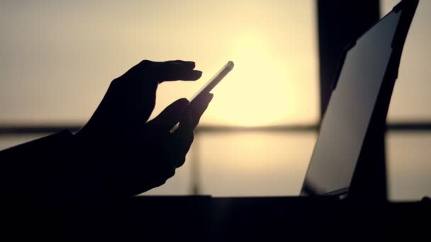 Volgende is donker silhouet van vrouwelijke handen, iets te typen in mobiele telefoon, laptop op tafel. tegen de achtergrond van een grote loket, bij zonsondergang, in de stralen van licht. Close-up — Stockvideo