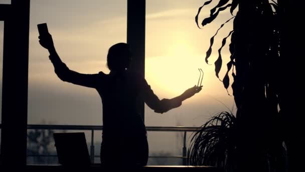 Temná silueta podnikatelka, tance s chytrým telefonem v ruce, na pozadí okna velké kanceláře, při západu slunce, v paprscích světla. obchodní žena slaví vítězství — Stock video