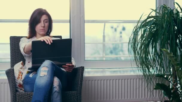 一个年轻的女人, 一个女孩, 一个穿白衬衫和牛仔裤的黑发女人, 坐在椅子上, 带着笔记本电脑。她在键盘上输入的东西, 看着显示器, 笔记本电脑屏幕。在一个大的背景下 — 图库视频影像