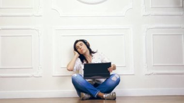 Genç kadın, kız, beyaz gömlek ve pantolon, bacaklar ile oturan yere dekoratif sıva ile beyaz duvar karşıya geçti kulaklık ve bir dizüstü bilgisayar kullanarak müzik dinleme esmer,