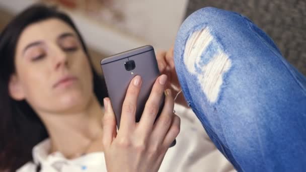 Młoda kobieta, Dziewczyna, Brunetka, biała koszula i jeansy, leżąc na kanapie, mała kanapa z smartphone. słuchanie muzyki, patrzy na ekran, bierze sobie selfie wideo, grymasy. niewyraźne tło — Wideo stockowe