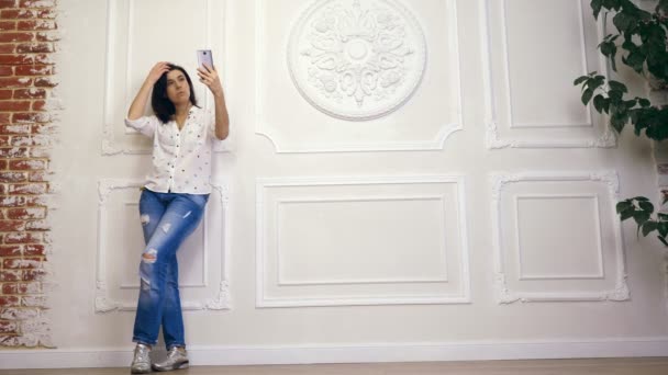 Genç kadın, kız, esmer, beyaz gömlek ve beyaz duvar smartphone ile dekoratif sıva ile arka plan üzerinde ayakta kot, tam uzunlukta portresi. ekranında görünüyor, selfie alır, — Stok video