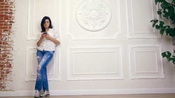 Portrait complet de jeune femme, fille, brune, en chemise blanche et jean, debout sur fond de mur blanc avec stuc décoratif, avec smartphone. regarde l'écran, en tapant quelque chose dans un — Video