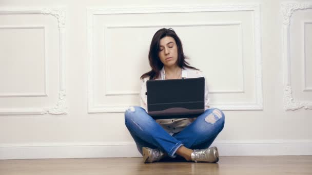 Pełna długość Portret młodej kobiety, dziewczyny, Brunetka, biała koszula i jeansy, praca na komputerze przenośnym, podczas gdy siedzi na podłodze z nogami skrzyżowanymi, na tle białej ściany z ozdobne stiuki, — Wideo stockowe