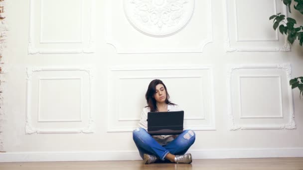 Volledige lengte portret van de jonge vrouw, meisje, brunette, in wit shirt en spijkerbroek, werken op laptopcomputer terwijl zittend op de vloer met de benen, op achtergrond van witte muur met decoratief stucwerk gekruist, — Stockvideo