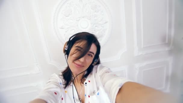 Portrét legrační mladá žena, dívka, brunetka, v bílé košili, sluchátka, ona poslouchá hudbu, střílí video selfie nad bílou stěnu s dekorativní štuky, dívka dělá legrační, hloupé tváře, — Stock video