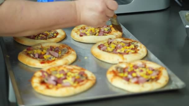 Primo piano, il processo di cottura di diverse mini pizza da pasta lievitata, con salsiccia e formaggio. Lo chef spruzza pizza con formaggio. processo di cottura panificio, in sala mensa, resturant . — Video Stock