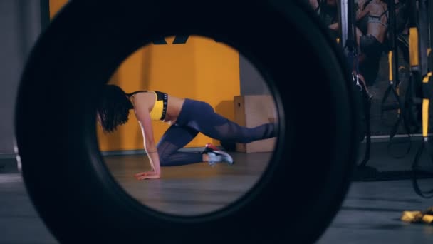 Mulher morena atraente em leggings, top amarelo e tênis executa exercícios em loops TRX, Correias no ginásio. um olhar através do grande pneu trator — Vídeo de Stock