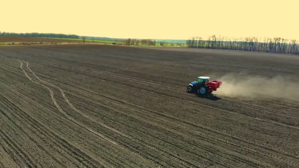 Aerial view of agricultural tractor on big wheels rides across the fertile farm field, spreading mineral fertilizers with seed drill, raises great dust on ploughed ground. Spring soil cultivation — Stock Video