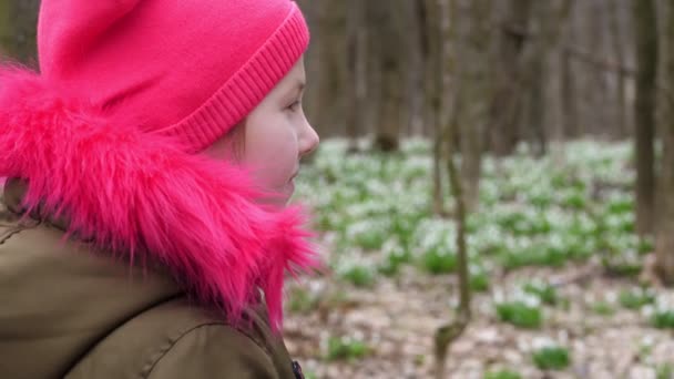 Portrait d'une adolescente portant un chapeau rose vif et une veste de couleur kaki avec une fourrure rose vif sur fond de gouttes de neige en fleurs, dans la forêt printanière — Video