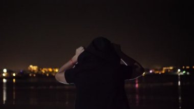 Gece, siyah kapüşonlu bir adam dans ediyor, arkadan manzara. suyun üzerinde parlayan şehrin ışıklarına karşı