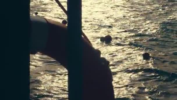 Primer plano, olas marinas al atardecer, en los rayos de luz solar suave. resplandor en el agua. contornos de una boya salvavidas que cuelga del pantone, muelle. los turistas nadan con máscaras para bucear en el mar . — Vídeo de stock