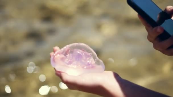 Közeli, kezében egy medúza, spárgasípok a mobil telefon, a háttérben a víz — Stock videók
